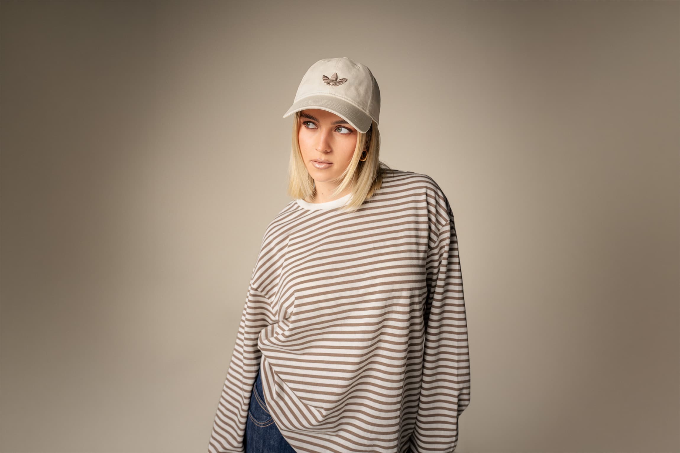 Frau in einem gestreiften Langarmshirt und einer beigen Mütze mit Blumenlogo steht vor einem neutralen Hintergrund und schaut zur Seite.