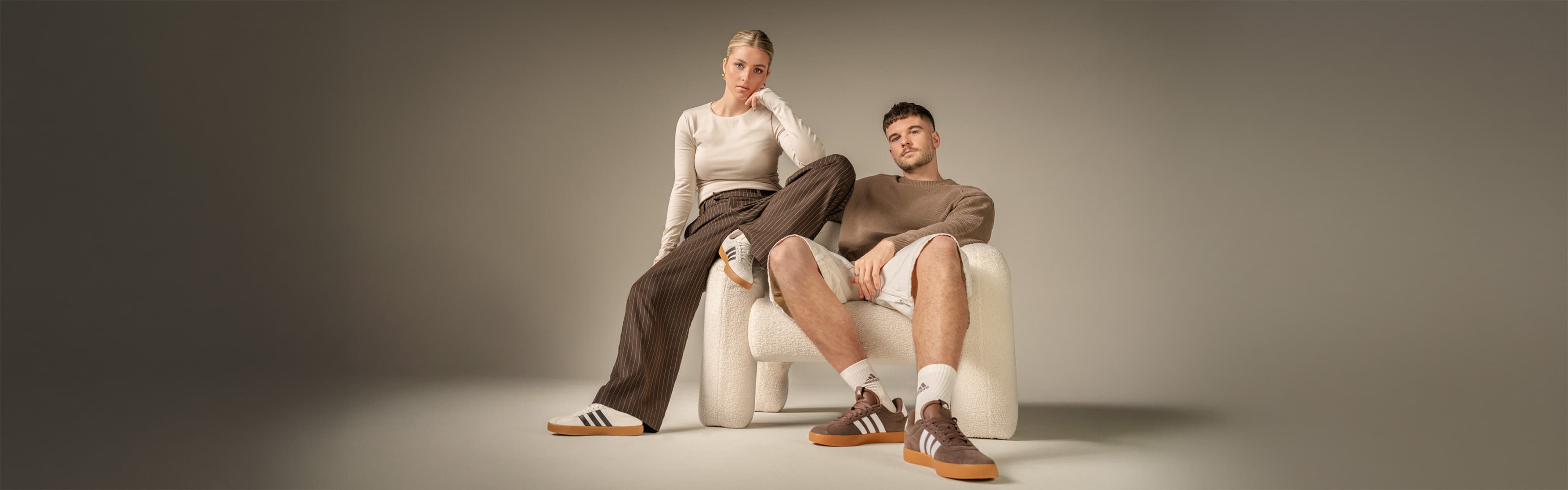 Eine Frau steht neben einem Mann, der auf einem weißen Stuhl sitzt. Sie trägt ein weißes Oberteil, eine braune Hose und Turnschuhe, während er einen braunen Pullover und Shorts trägt.