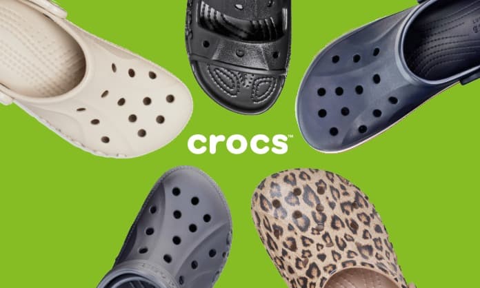 Fünf Crocs-Clogs in verschiedenen Farben und Mustern, darunter Beige, Schwarz, Marineblau, Grau und Leopardenmuster, die auf grünem Hintergrund kreisförmig angeordnet sind.