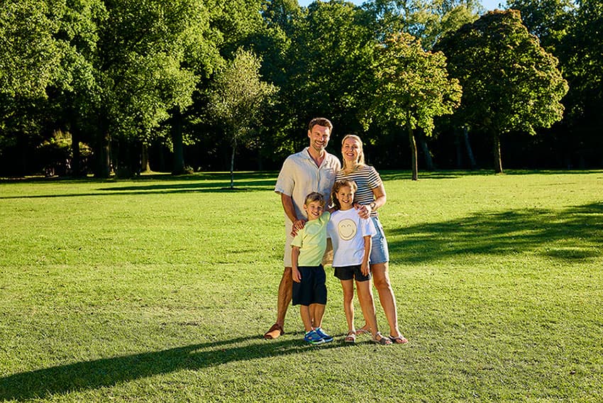 Eine vierköpfige Familie steht auf saftigem grünem Gras in einem sonnigen Park, mit Bäumen im Hintergrund, lächelt und genießt einen strahlenden, sonnigen Tag.